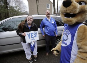 In this Tuesday, March 15, 2016 photo, Maya Wolf, 17, left, holds a sign to her mother, not shown, from the driveway of their home in Franklin, Mass., as she is visited by Wheaton College President Dennis Hanno, center, and the school's mascot. Wolf was presented with an acceptance letter from Wheaton College officials during the visit. More colleges have started to hand-deliver portions of their acceptance letters to students. (AP Photo/Steven Senne)