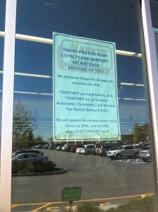 A sign on the door of the Sagamore Market Basket welcomes back customers. The glass shows the reflection of the full parking lot.